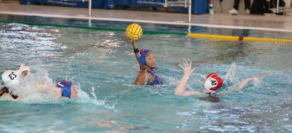 A water polo player holds up a yellow ball while other players splash around her.